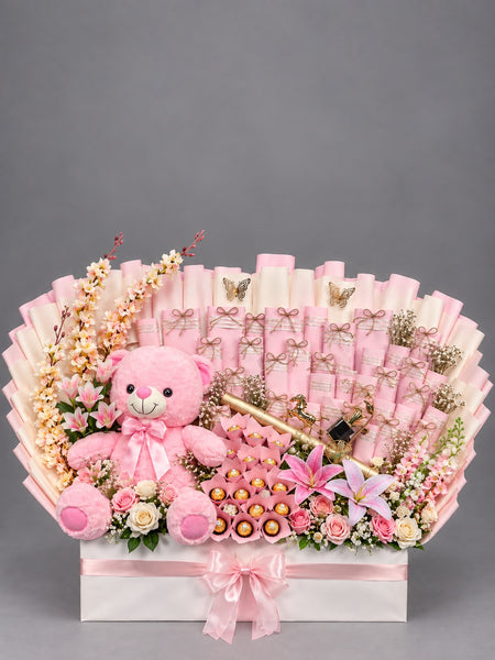 A pink-themed gift basket arrangement featuring a plush pink teddy bear, rolled pink paper fan elements, pink flowers, chocolates, and decorative gold accents, presented on a white box against a gray studio background.