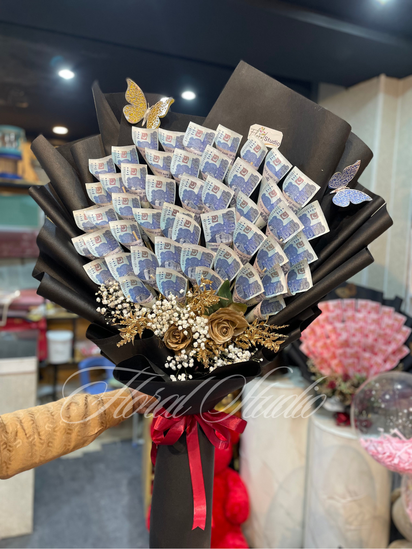 A bouquet made of folded Pakistani currency notes, artificial golden roses, white baby's breath, and golden foliage, wrapped in black paper with a red satin ribbon.