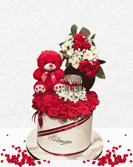 Floral arrangement with red roses, a teddy bear, and a small cake on a white background.