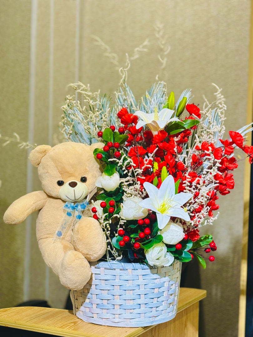 A teddy bear sitting next to a basket of red and white flowers.