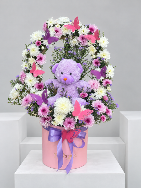 Lavender teddy bear in a wreath of pink and white chrysanthemums with butterflies in a pink box