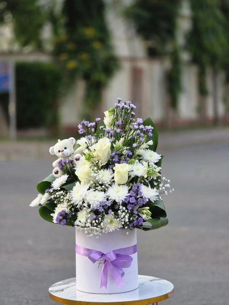 Floral arrangement in a white box with a purple ribbon and a teddy bear, set against a blurred outdoor background.