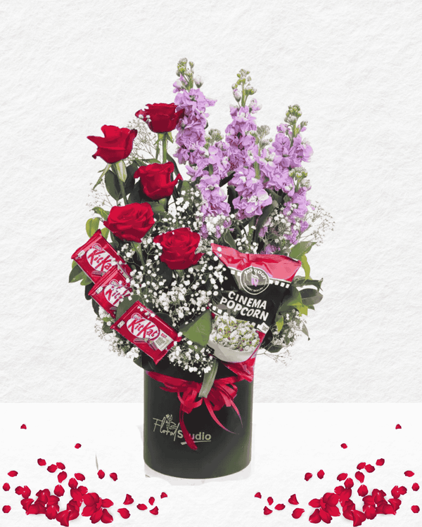 Bouquet of red roses and purple flowers with candy bars and popcorn packaging in a black container on a white background.