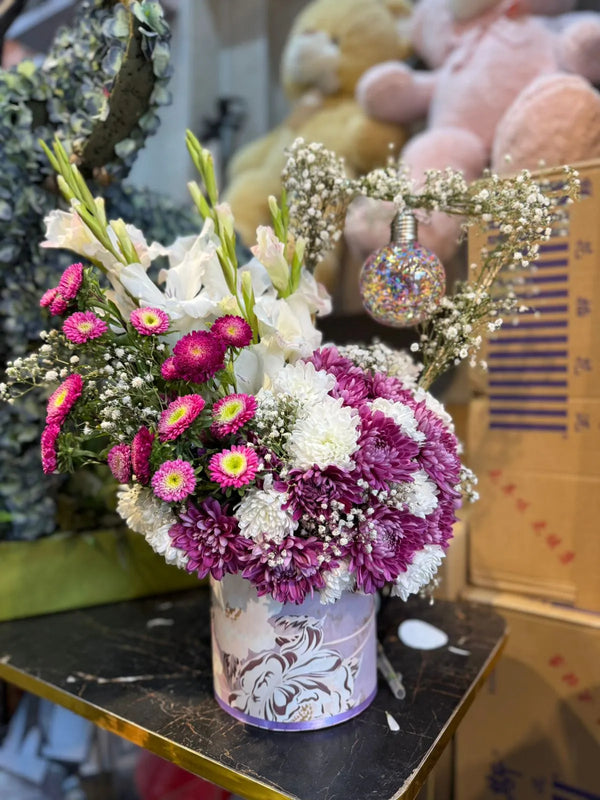 Floral arrangement in a decorative pot on a table with plush toys in the background