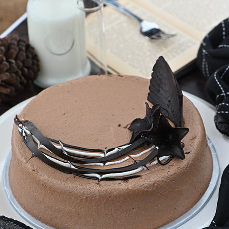A chocolate fudge cake with a decorative butterfly and pine cone on top, set on a plate with a candle and pine cones in the background.