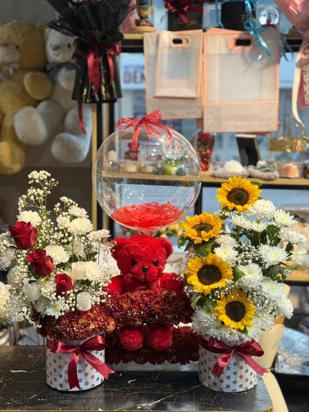 Decorative setup with flowers, a teddy bear, and a transparent bubble containing red roses on a table.