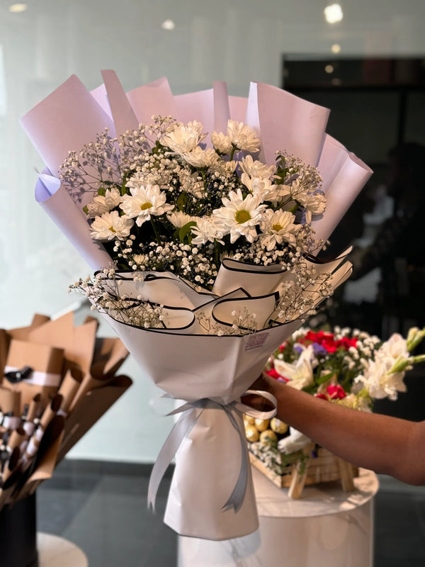 Bouquet of flowers wrapped in white paper with a silver ribbon, set against a blurred indoor background.
