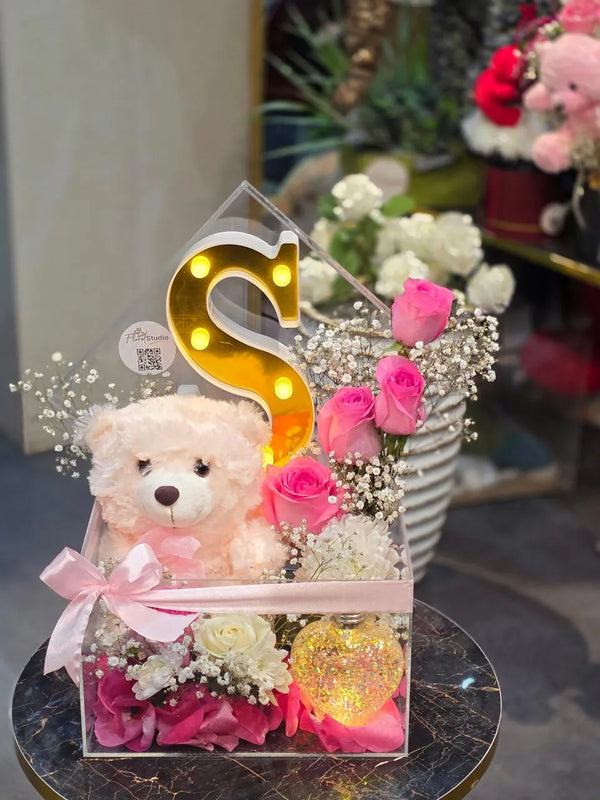 Teddy bear with flowers and a letter 'S' in a decorative box.
