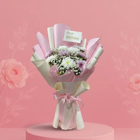 Bouquet of flowers wrapped in white paper with a 'Happy Birthday' card on a pink platform against a pink background.