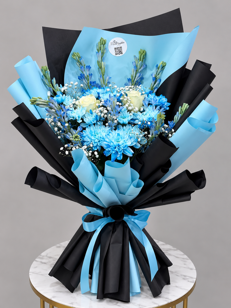 A bouquet of blue and white flowers wrapped in black and light blue paper, displayed on a marble-topped table against a gray background.