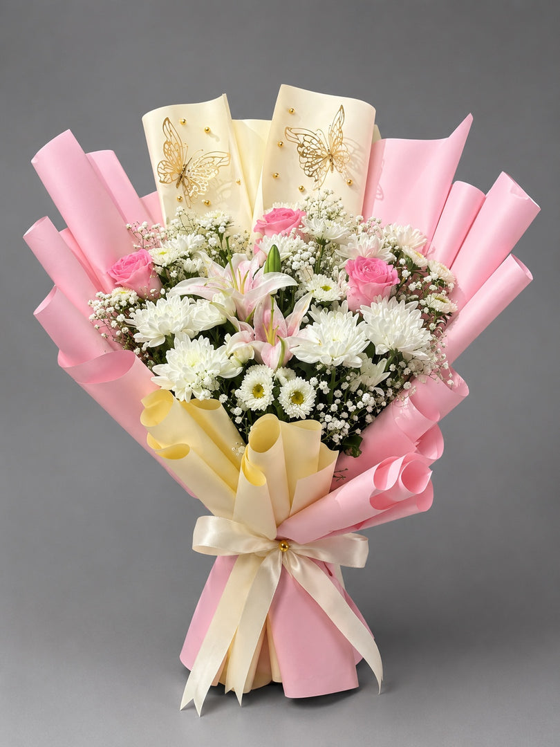 A floral bouquet wrapped in pink and cream paper with gold butterfly designs, featuring white and pink flowers, including roses, lilies, and daisies, tied with a cream ribbon against a solid gray background.
