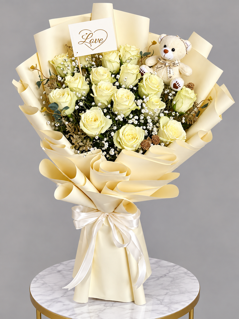 A bouquet of pale yellow roses wrapped in cream paper with a small teddy bear and a card reading 'Love' on a marble-topped table against a gray background.