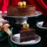A slice of Ferrero Rocher cake on a plate with the rest of the cake in the background, decorated with hazelnut cream and a gold-wrapped Ferrero Rocher on top.