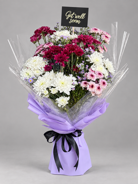 A vibrant mixed flower bouquet featuring white, deep magenta, and light pink chrysanthemums, wrapped in clear cellophane with a lavender paper sleeve tied with a black ribbon. A small black card at the top reads 'Get well soon'.