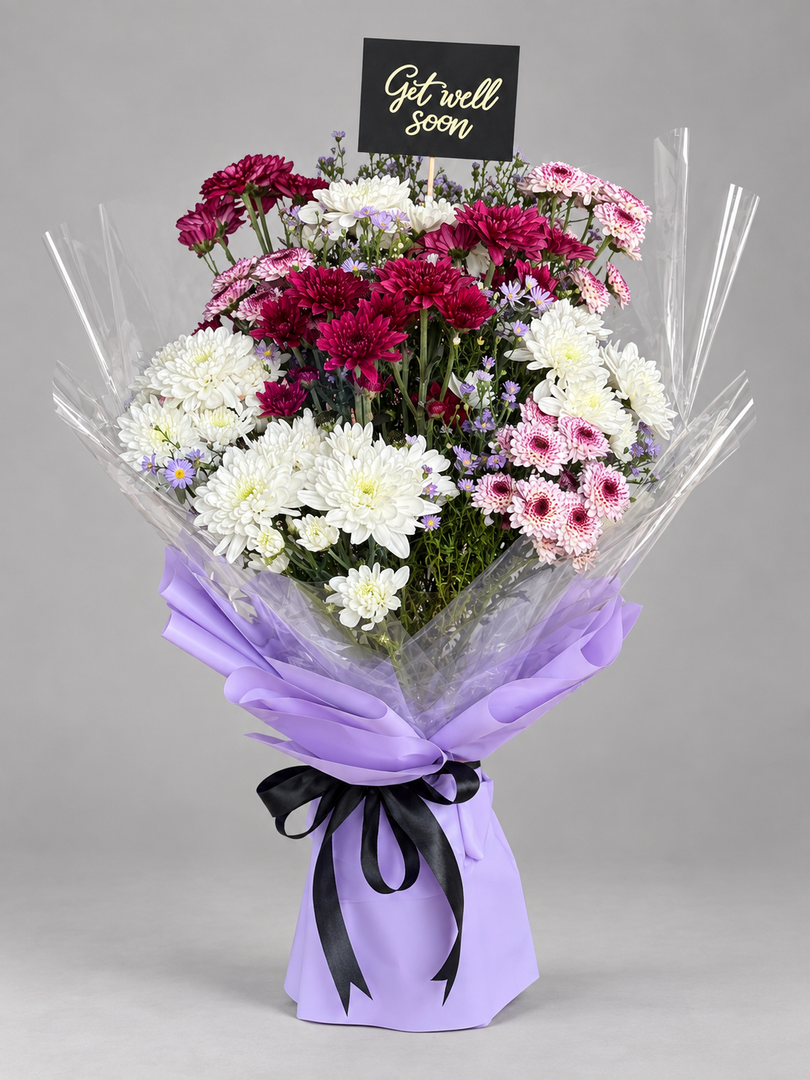 A vibrant mixed flower bouquet featuring white, deep magenta, and light pink chrysanthemums, wrapped in clear cellophane with a lavender paper sleeve tied with a black ribbon. A small black card at the top reads 'Get well soon'.