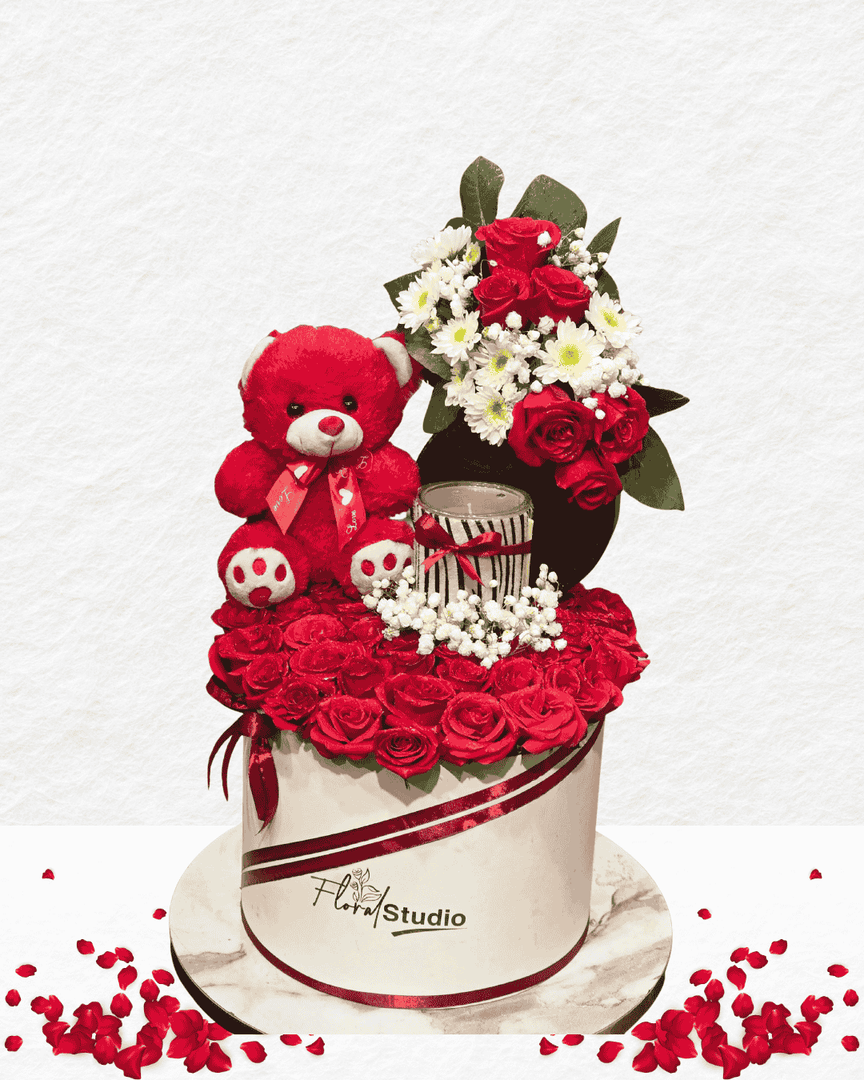 Floral arrangement with red roses, a teddy bear, and a small cake on a white background.