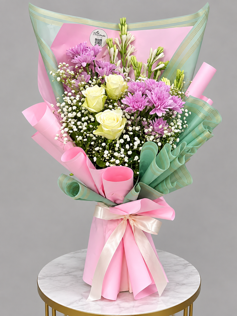 A vibrant floral bouquet featuring yellow roses, purple chrysanthemums, and white baby's breath, wrapped in pink and mint green paper with a pink ribbon, displayed on a marble-topped table against a neutral gray background.