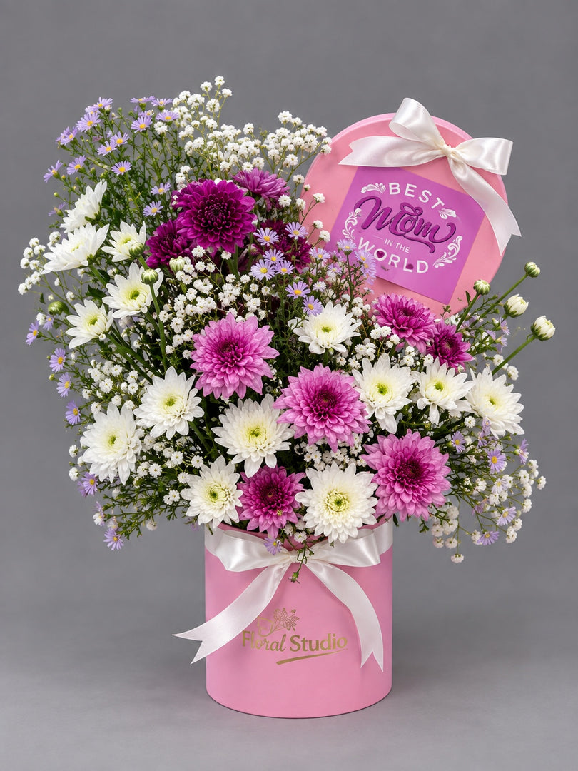 A floral gift arrangement in a pink cylindrical box with a pink heart-shaped card reading 'Best Mom in the World.' The bouquet includes white and magenta chrysanthemums, small white and lavender baby's breath, and white ribbon bows.