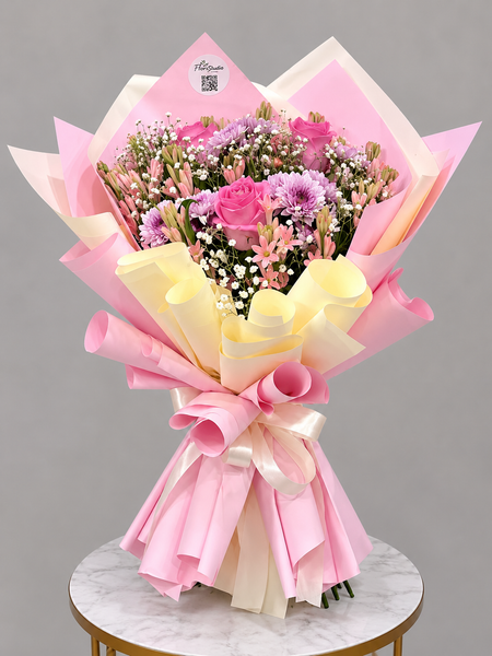 A vibrant flower bouquet wrapped in pink and pale yellow paper, featuring pink roses, purple chrysanthemums, and white baby's breath, displayed on a marble-topped stool against a neutral gray background.