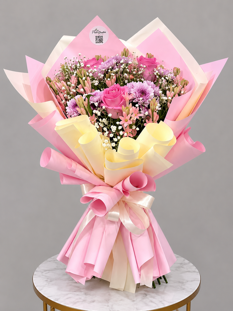 A vibrant flower bouquet wrapped in pink and pale yellow paper, featuring pink roses, purple chrysanthemums, and white baby's breath, displayed on a marble-topped stool against a neutral gray background.