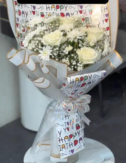 Bouquet of white flowers wrapped in 'Happy Birthday' paper on a gray surface. Send flower to Pakistan.