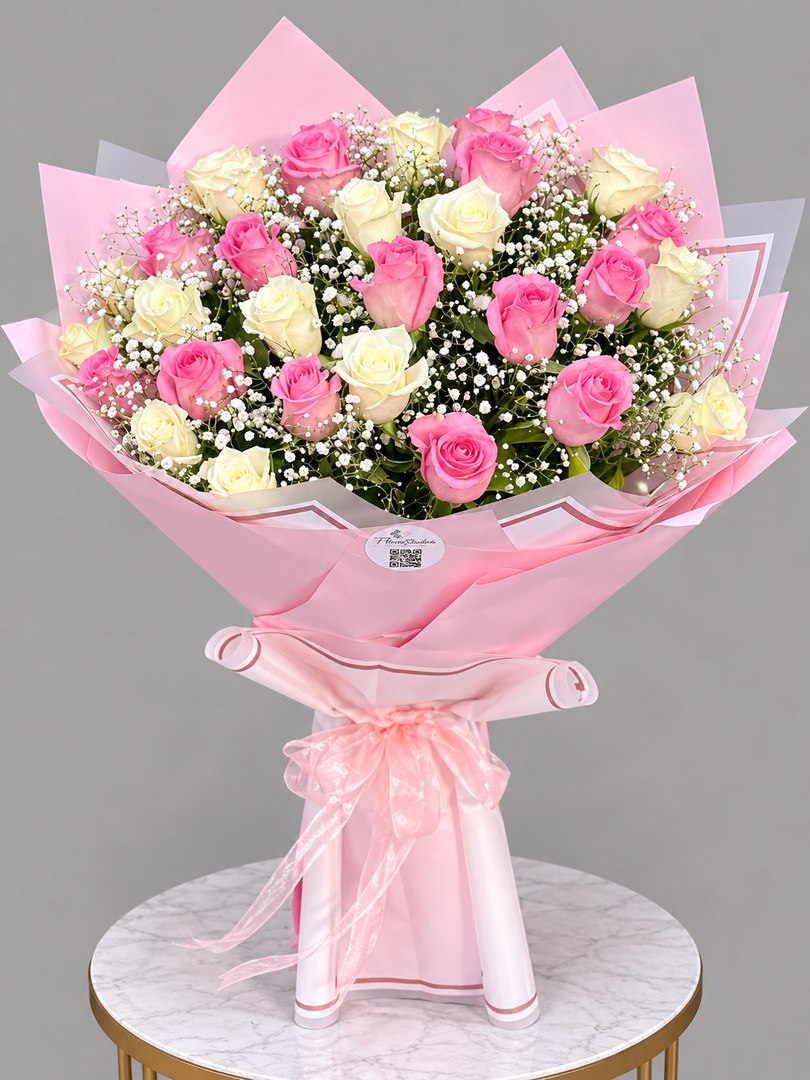 A large bouquet of pink and white roses with green foliage and baby's breath, wrapped in pink tissue paper and tied with a pink ribbon, displayed on a marble-topped table against a gray background.