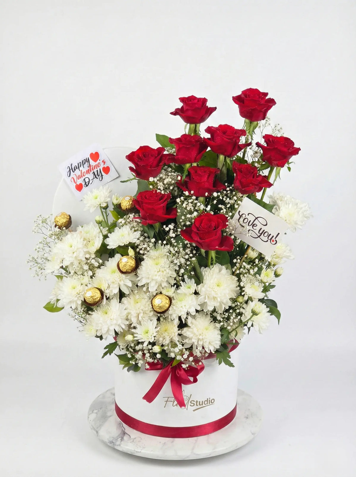 Floral arrangement with red roses and white flowers in a box, featuring a visible brand name on a white background.