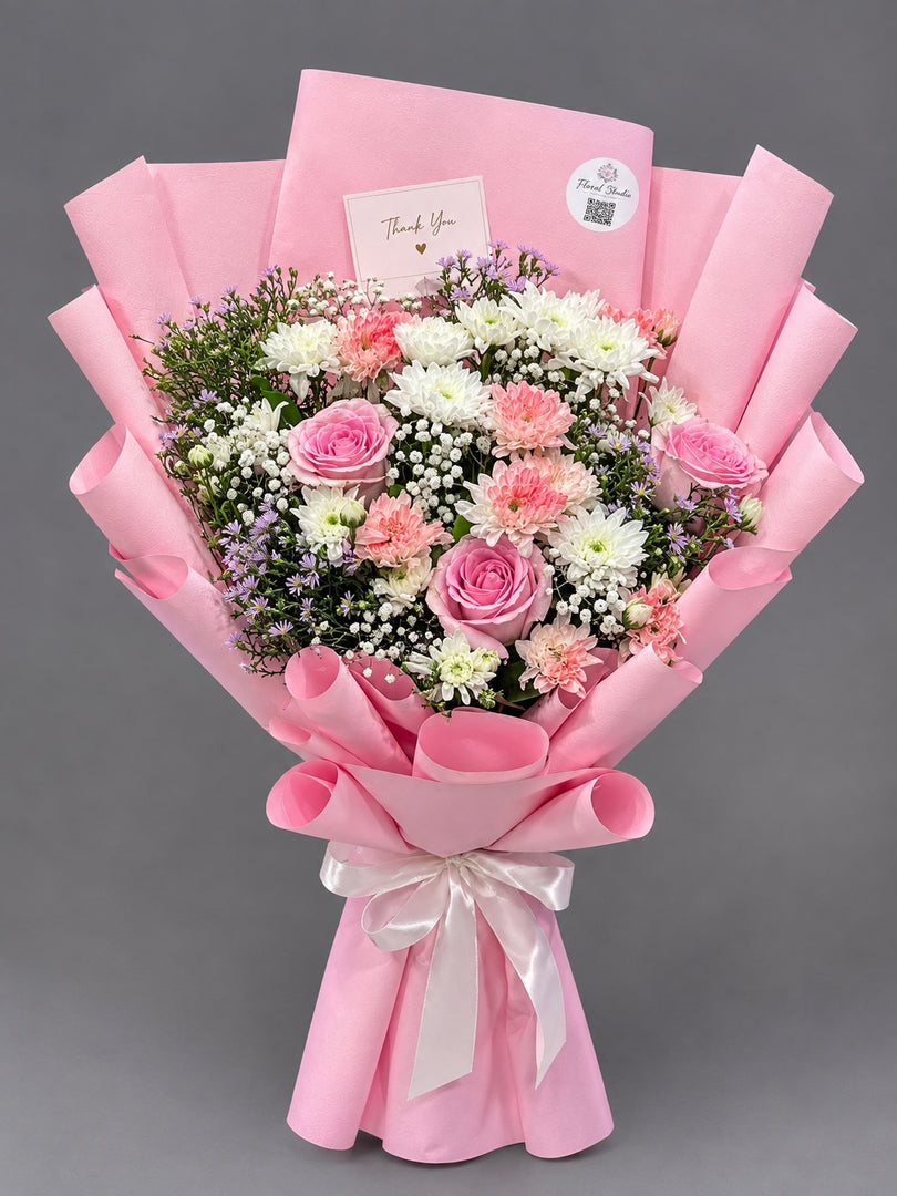 A pink wrapped flower bouquet with pink, white, and light pink roses, mixed with white baby's breath and green foliage, presented with a pink card and envelope on a gray background.