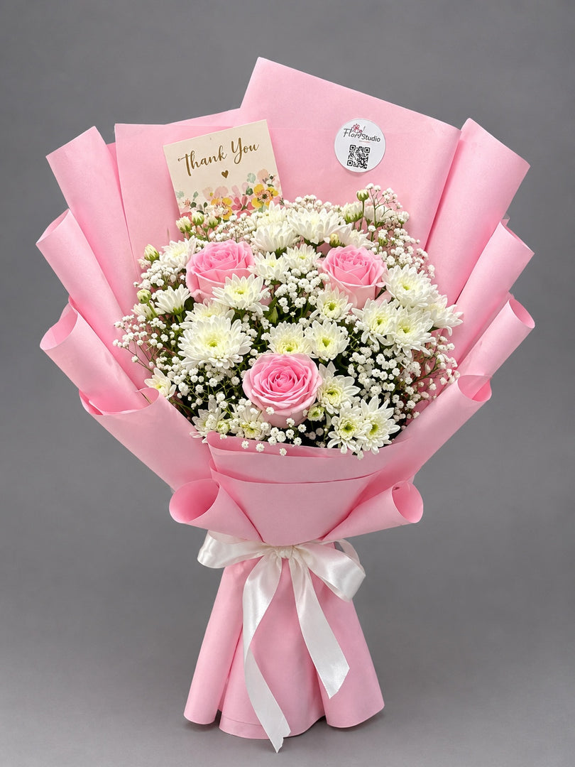 A floral bouquet wrapped in pink paper with a white ribbon, featuring pink roses, white daisies, and baby's breath, with a thank you card and round sticker on a gray background.