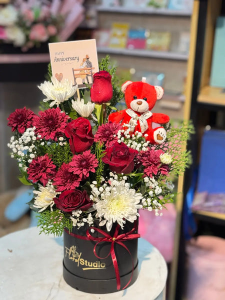 Floral arrangement with red roses and a teddy bear in a black box, featuring a card on top. Send flowers to lahore.