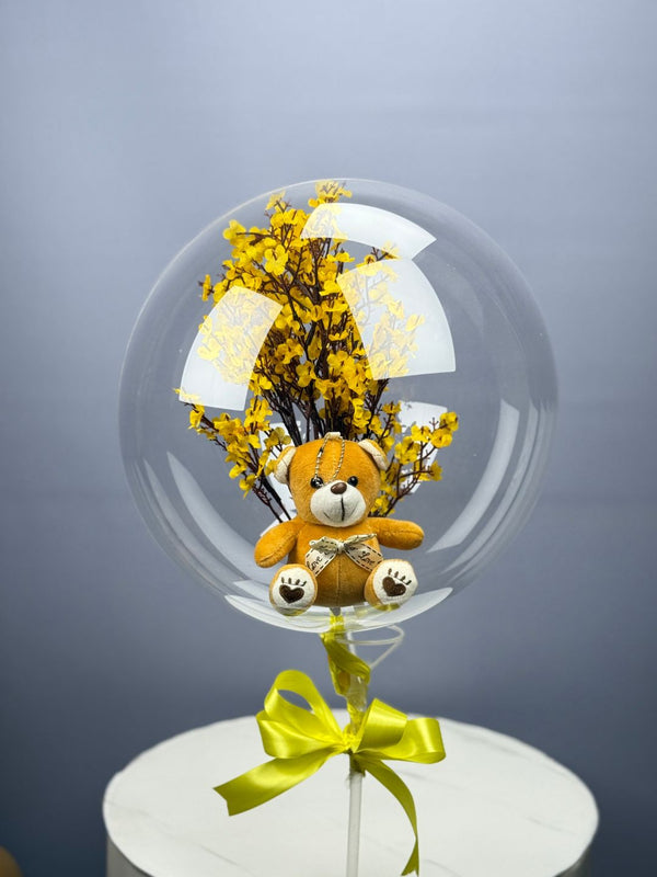 A clear balloon with a teddy bear and yellow flowers inside, tied with a yellow ribbon on a white stand.