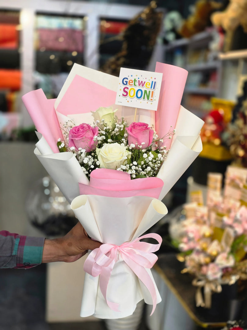 Bouquet of flowers with pink ribbons and a 'Get well soon!' card held by a person in a store setting.