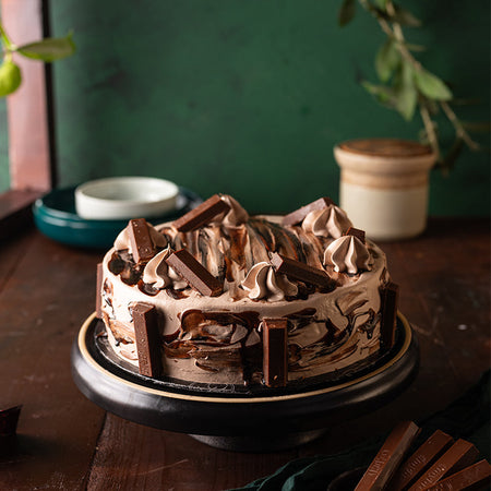 A chocolate cake with layers of chocolate cream and KitKat pieces on a round serving platter, with a plant in the background.
