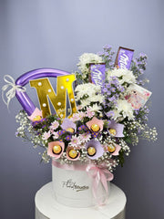 Floral arrangement with 'M' balloons and chocolate bars in a white box on a gray background. Send flowers to Lahore.