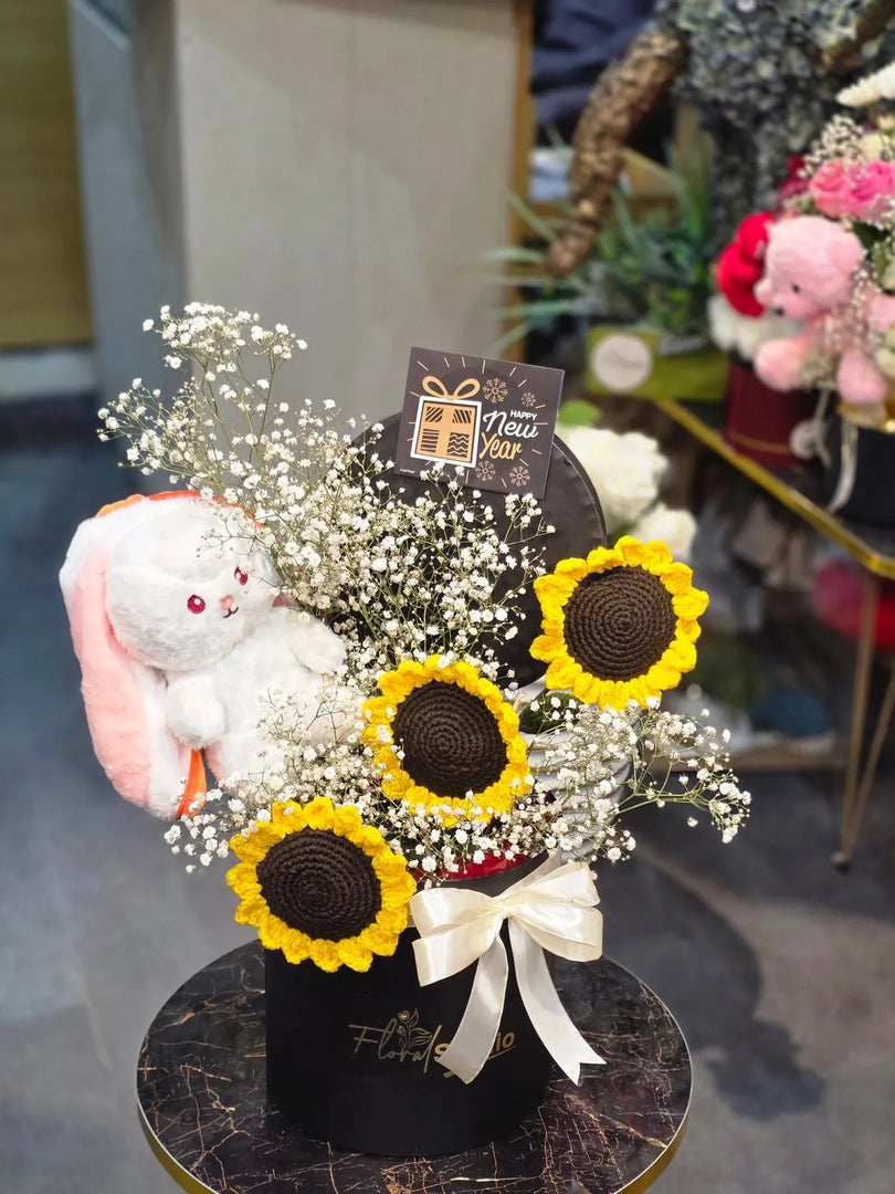 Floral arrangement with sunflowers, a plush toy, and a card on a black base.