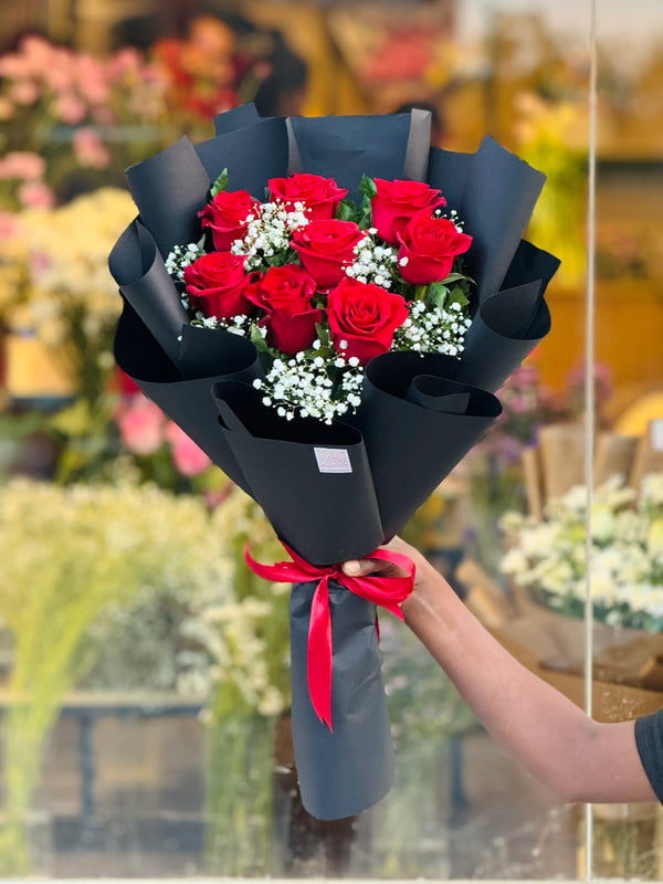 Bouquet of red roses with a black wrap and red ribbon held by a person in a blurred floral shop setting. Send flowers to lahore