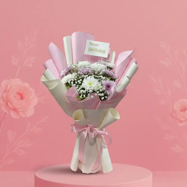 Bouquet of flowers wrapped in white paper with a 'Happy Birthday' card on a pink platform against a pink background.
