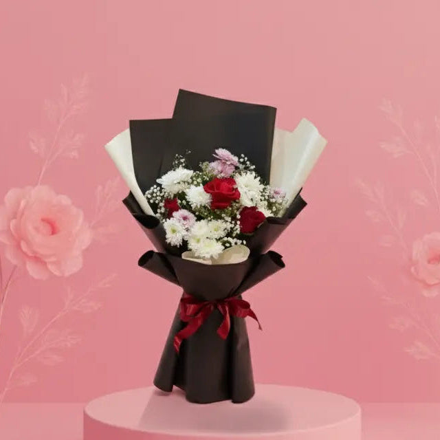Bouquet of flowers wrapped in black paper with a red ribbon on a pink platform against a pink background.
