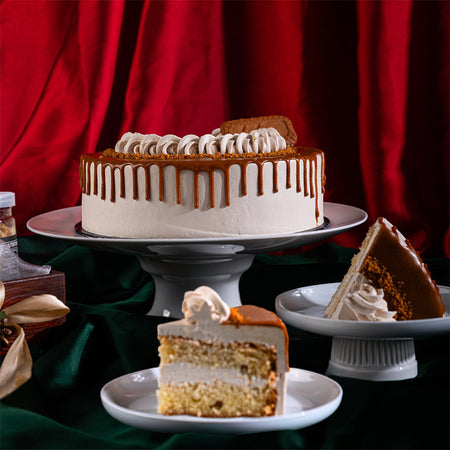 A sliced cake on a plate with a slice served separately, showing layers of moisture and cream, with a decorative drizzle on top, placed on a table with a red curtain in the background.