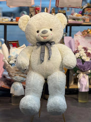 Large white teddy bear with a bow tie in a store setting with flowers and gift bags.