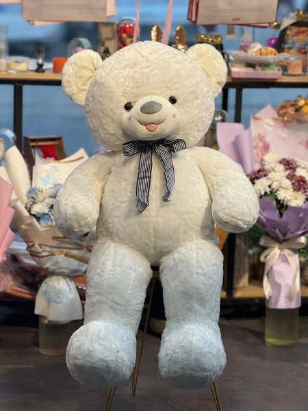 Large white teddy bear with a bow tie in a store setting with flowers and gift bags.