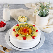 A vanilla sponge cake layered with fruit and whipped cream, served on a plate with a bottle of milk and a bowl of pineapples in the background.