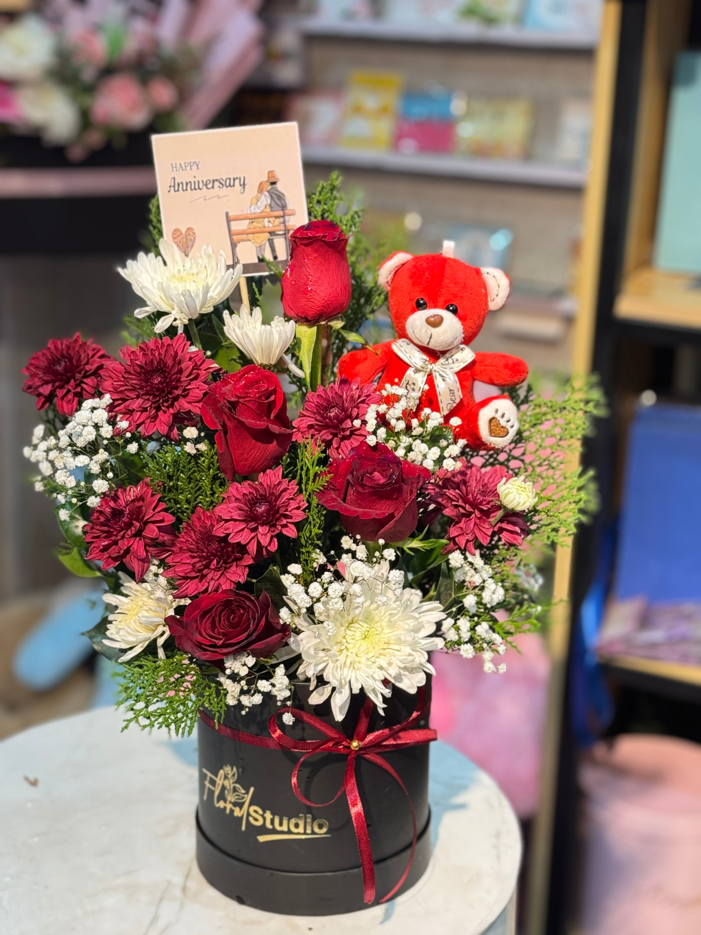 Anniversary Bliss floral arrangement featuring red roses, white chrysanthemums, maroon mums, baby’s breath, a red teddy bear, and an anniversary card in a black luxury box with a red ribbon.