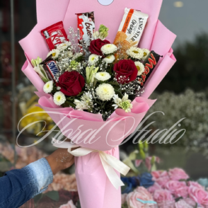 Bloom Bites bouquet featuring fresh red roses, white chrysanthemums, and chocolates including KitKat, Snickers, Mars, wrapped in pink paper with a ribbon tie.