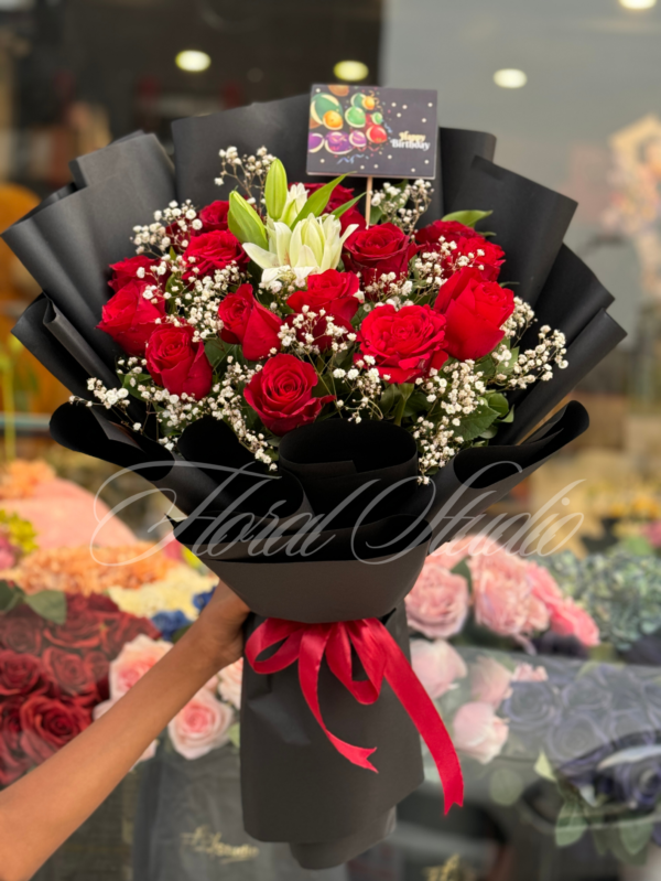 Crimson Charm bouquet with red roses, white lilies, and baby's breath wrapped in black paper with a red ribbon – ideal for special occasions.