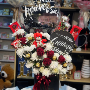 Anniversary Charm Floral Box featuring fresh red roses, white chrysanthemums, Ferrero Rocher chocolates, a teddy bear, and a personalized Happy Anniversary balloon in a sleek black box with red ribbon. Perfect for anniversary celebrations and available for online flowers delivery in Lahore.