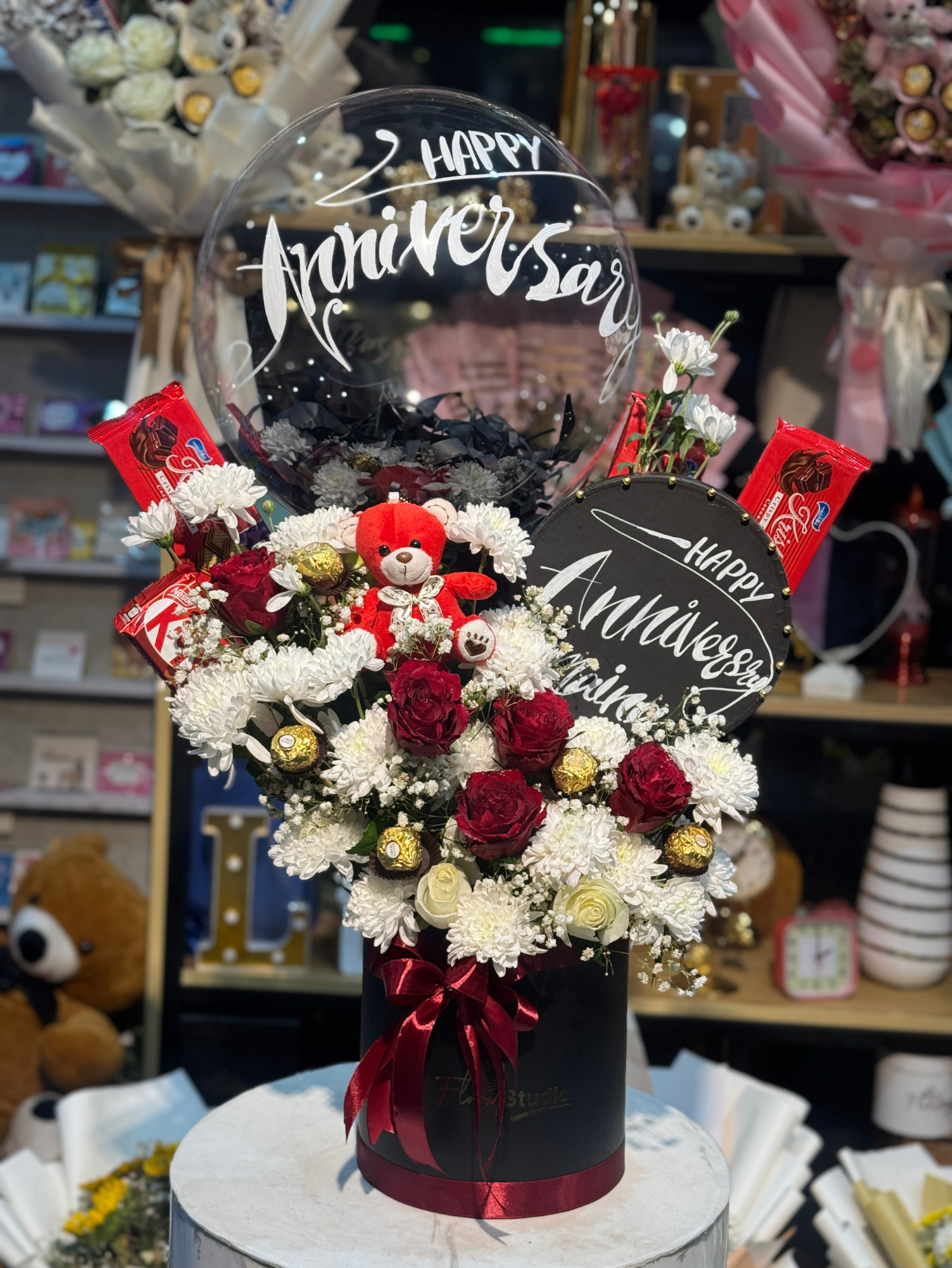 Anniversary Charm Floral Box featuring fresh red roses, white chrysanthemums, Ferrero Rocher chocolates, a teddy bear, and a personalized Happy Anniversary balloon in a sleek black box with red ribbon. Perfect for anniversary celebrations and available for online flowers delivery in Lahore.