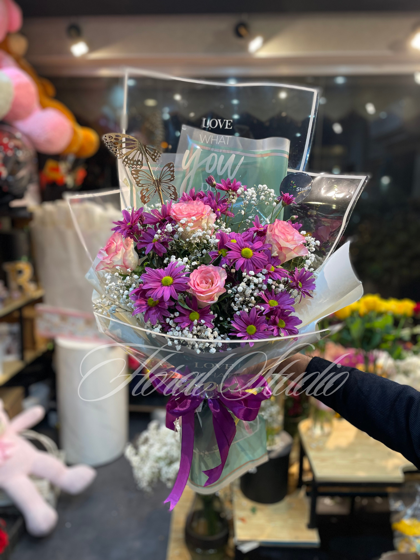 Vibrant flower bouquet featuring purple chrysanthemums, pink roses, white baby’s breath, and butterfly accents wrapped in transparent luxury packaging with a purple ribbon.