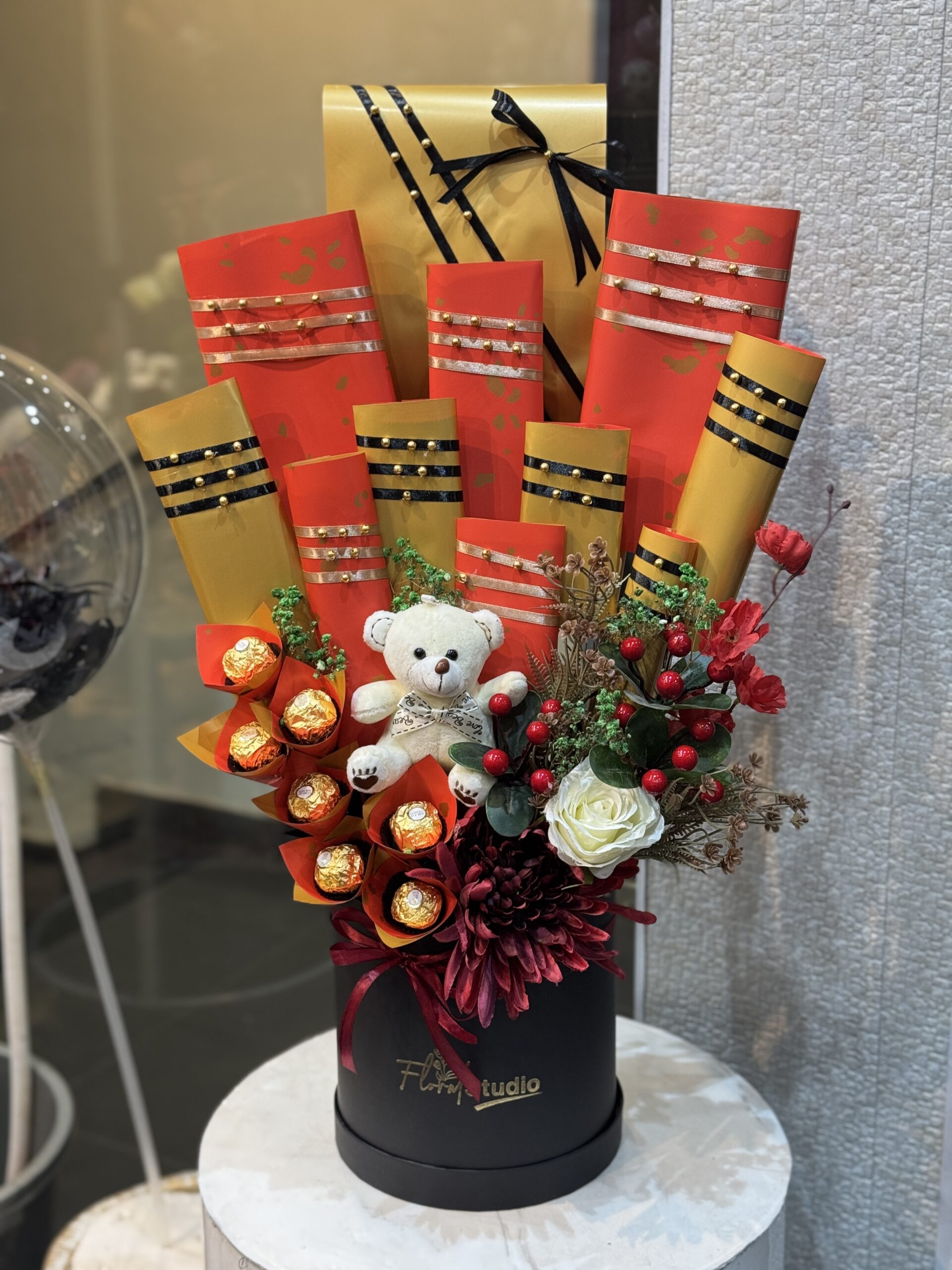 Golden Bliss Basket featuring Ferrero Rocher chocolates, a plush teddy bear, fresh flowers, and elegant gold and red packaging in a black box, perfect for gifting and available for online flower delivery in Lahore.