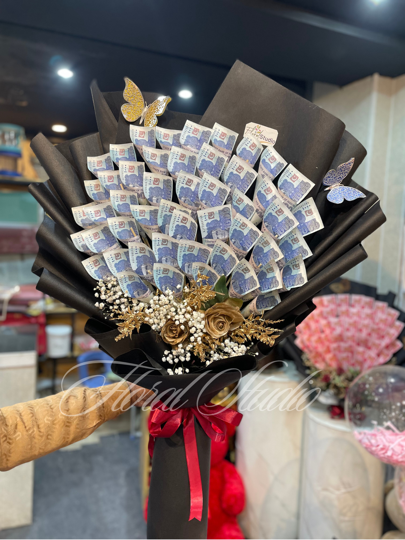 An opulent bouquet featuring carefully folded Pakistani currency notes, golden artificial roses, white baby's breath, and shimmering golden foliage, wrapped in elegant black paper and tied with a red satin ribbon, perfect for special celebrations and gifting.